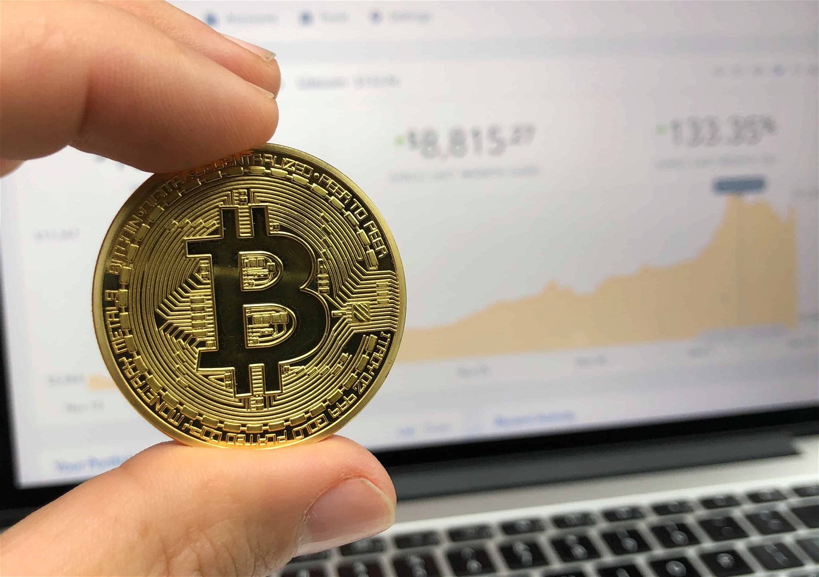 Een hand houdt een muntstuk met een 'B', verwijzend naar Bitcoin, tussen wijsvinger en duim met op de achtergrond het scherm van een pc waarop beurskoersen zijn afgebeeld.