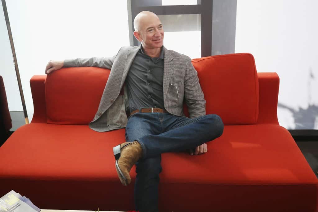 Jeff Bezos zit in kostuum ontspannen met gekruiste benen op een oranje zetel, terwijl hij één arm over de rugleuning legt en glimlacht.