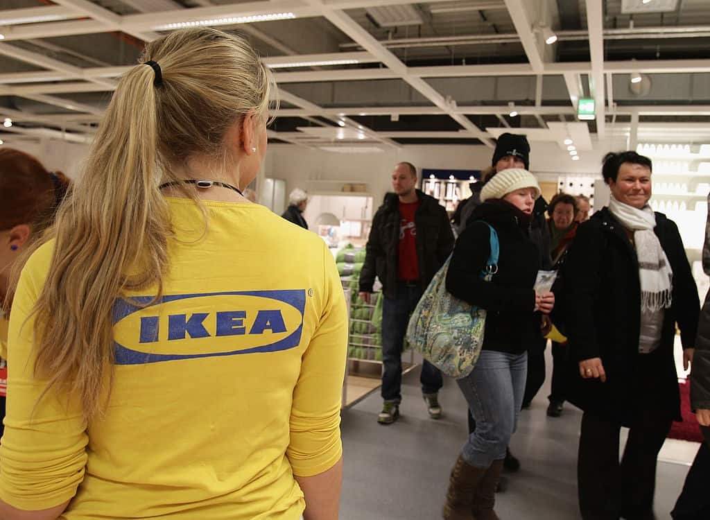 Een medewerkster van IKEA met blond haar en een geel t-shirt met het winkellogo kijkt toe terwijl klanten voorbijlopen.