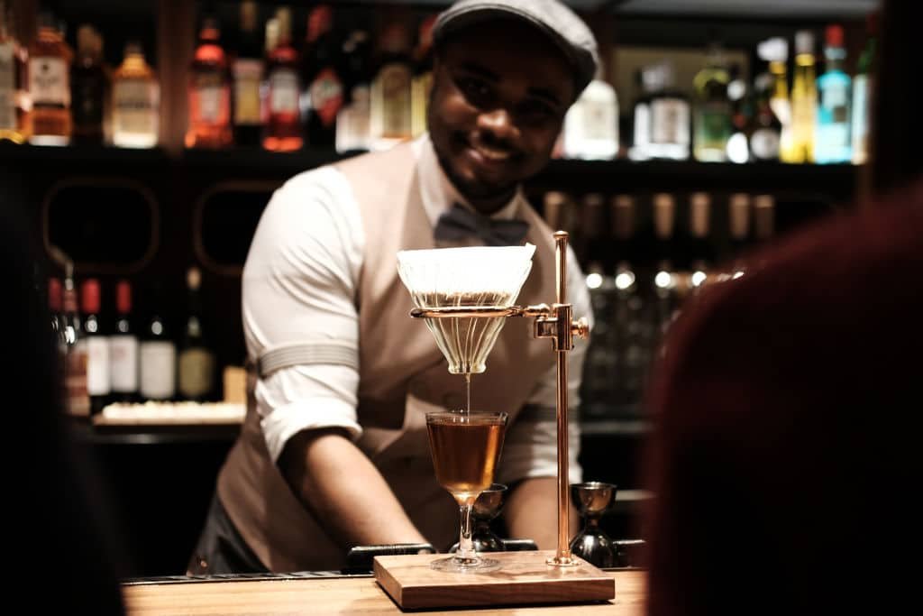 Een zwarte man lacht terwijl uit een koffiefilter vloeistof in een glas stroomt.
