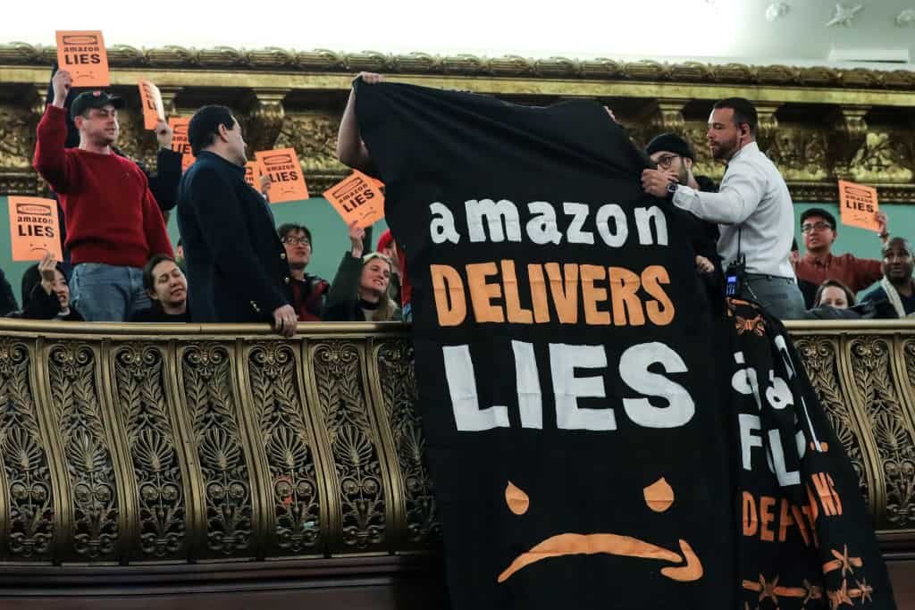 Medewerkers van Amazon houden een oranje bord vast en laten een doek naar beneden vallen met de tekst: "Amazon levert leugens."