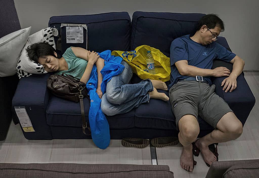 Een vrouw slaapt liggen op de linkerkant van een blauwe IKEA-zetel waar prijskaartjes aanhangen. Naast haar slaapt een man al zittend.