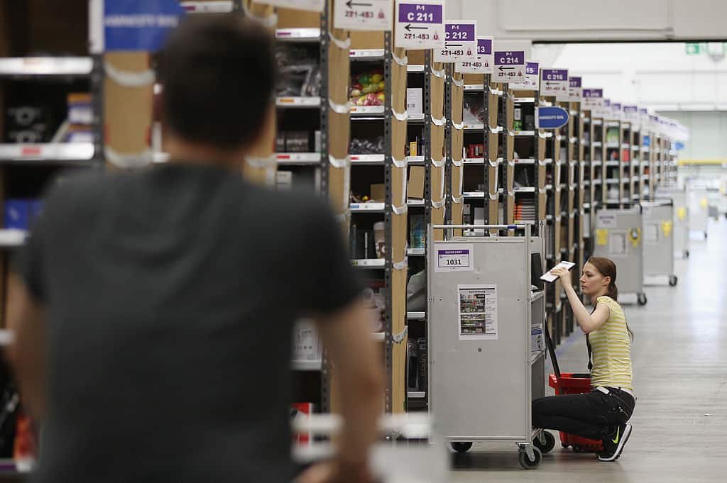 De schim van een man in een zwart shirt op de voorgrond, terwijl voor hem een jonge vrouw in een geel shirt op de grond zit aan talrijke gangen met items in een Amazon-magazijn.
