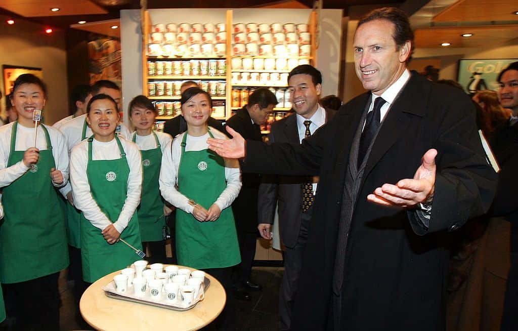 Vrouwen in een groene schort van Starbucks en een man met open armen staan rond een tafel met kopjes.
