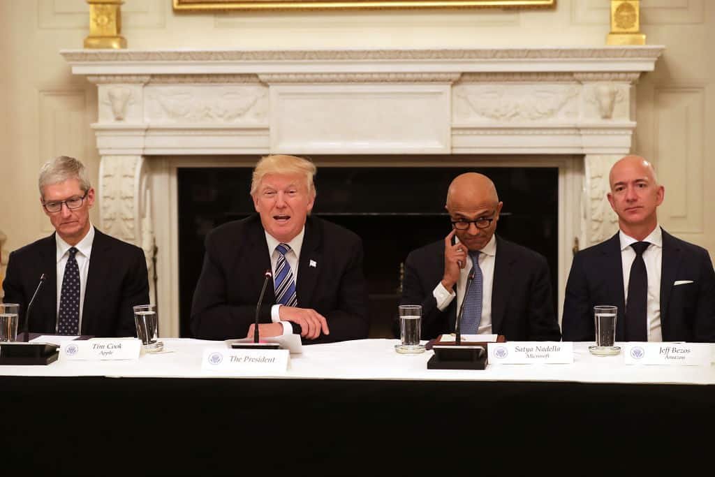Vier mensen zitten naast elkaar: Donald Trump is de tweede en spreekt, Jeff Bezos is de vierde in de rij en luistert met strak gezicht.