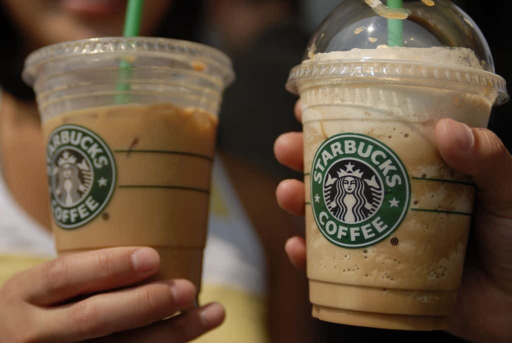 Twee handen houden een transparante beker van Starbucks gevuld met koffie vast.