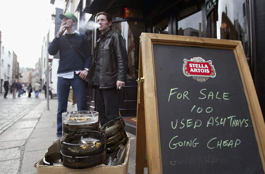 Asbakken staan voor de deur van een pub te koop, met op de achtergrond twee mannen die een sigaret roken.