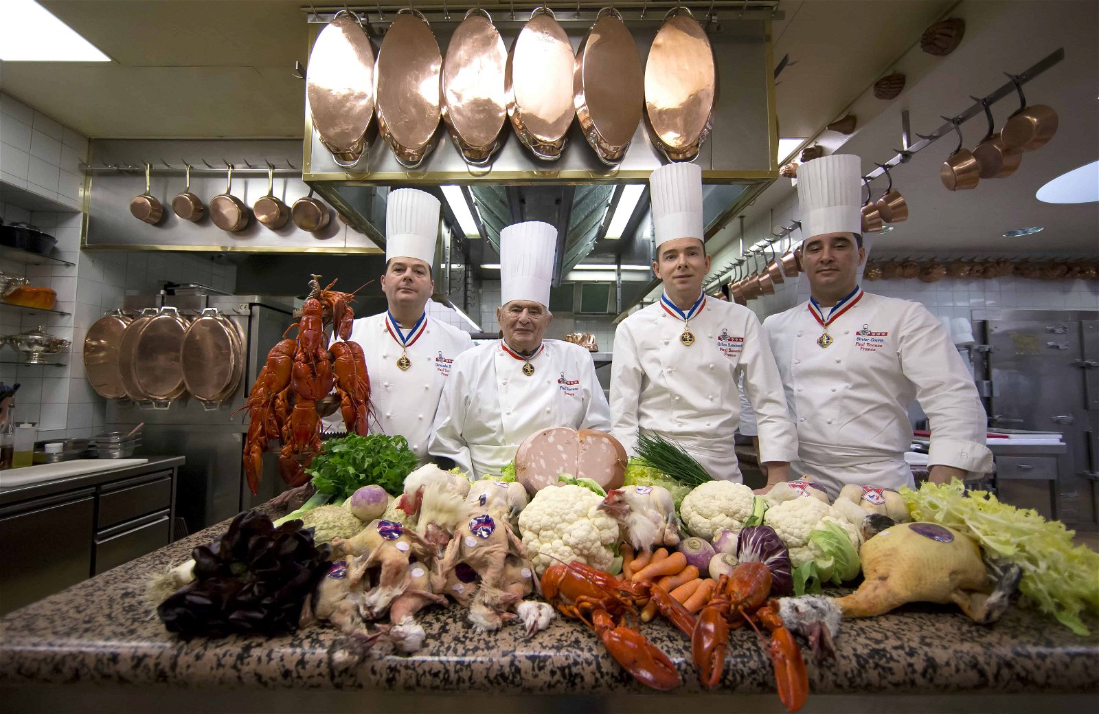 Vier koks in wit pak met hoge hoed poseren achter een kreeft en andere voedsel in de keuken.