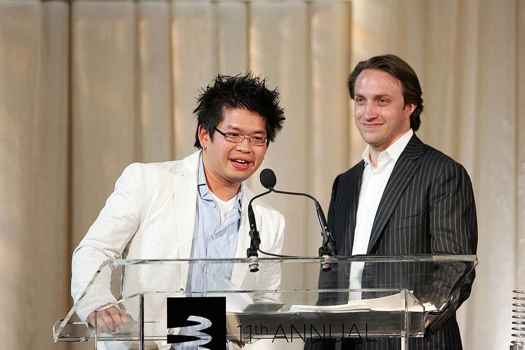 YouTube-oprichter Steve Chen praat in een wit kostuum voor een tafel met microfoon. Achter hem kijkt een man glimlachend toe.