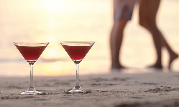 Two cocktails on the beach with couple kissing in the background, concept about romantic honeymoon in tropics